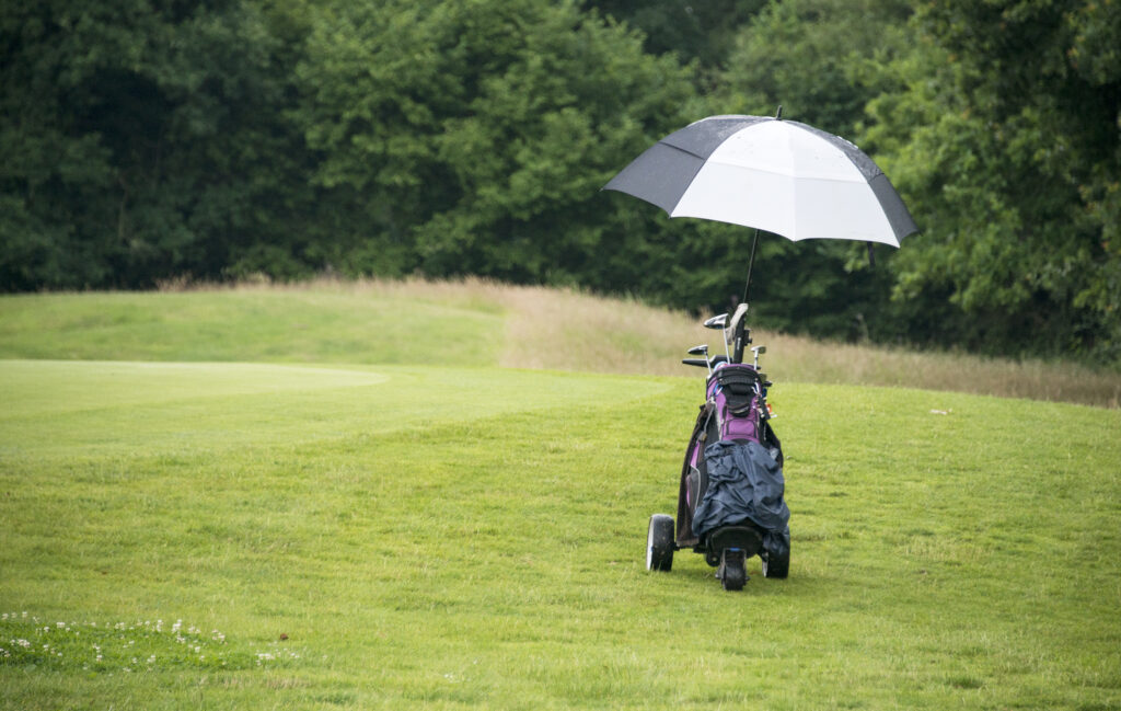 A golf bag with an umbrella on a pull cart sits next to a green during a light rain. 