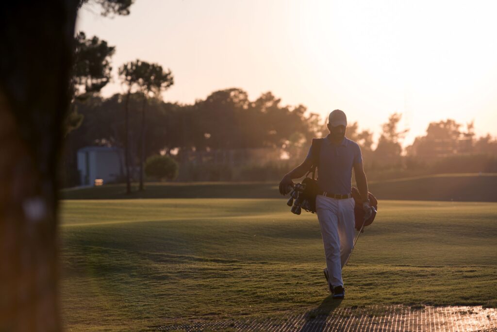 A golfer walks a golf course around sunrise carrying his clubs. 