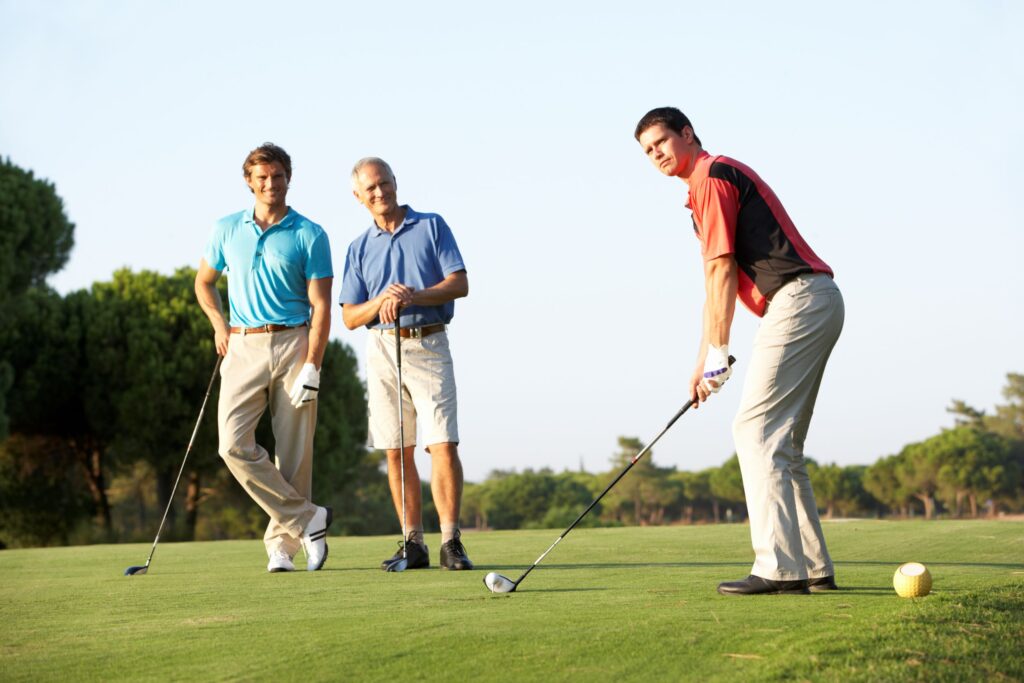 A golfer prepares to hit a golf shot while two other golfers watch. 