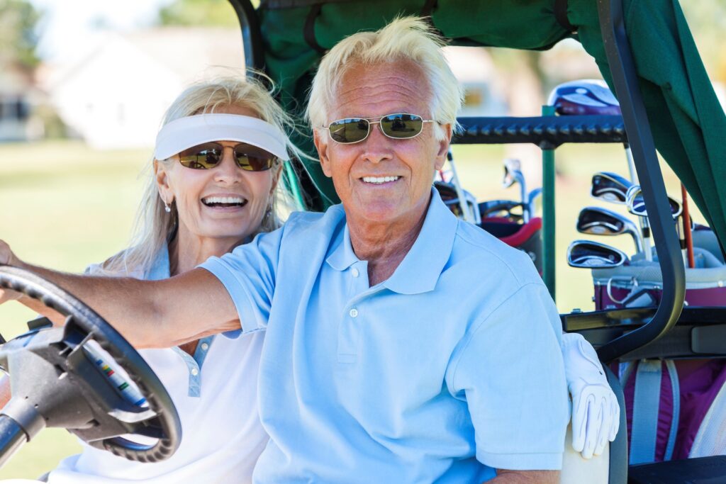 A senior couple riding in a golf cart with their golf clubs.