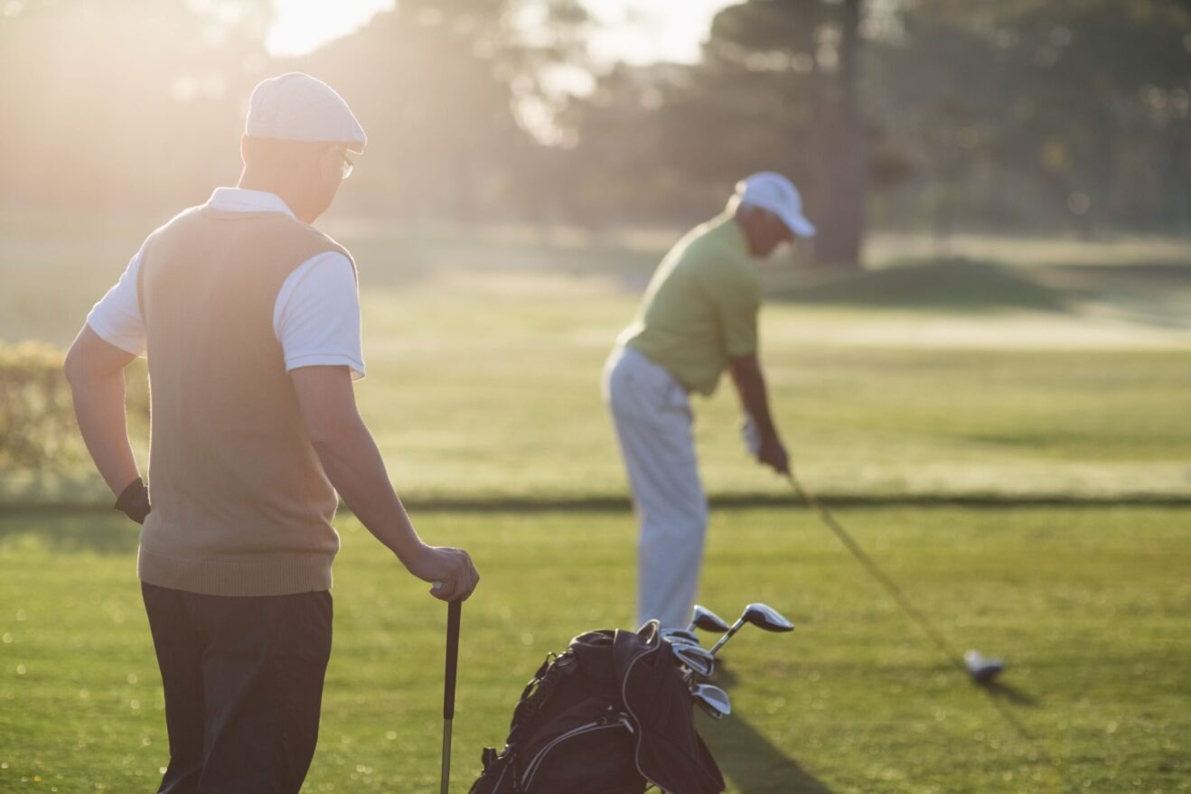 A golfer watches as their playing partner hits a drive on a golf course.