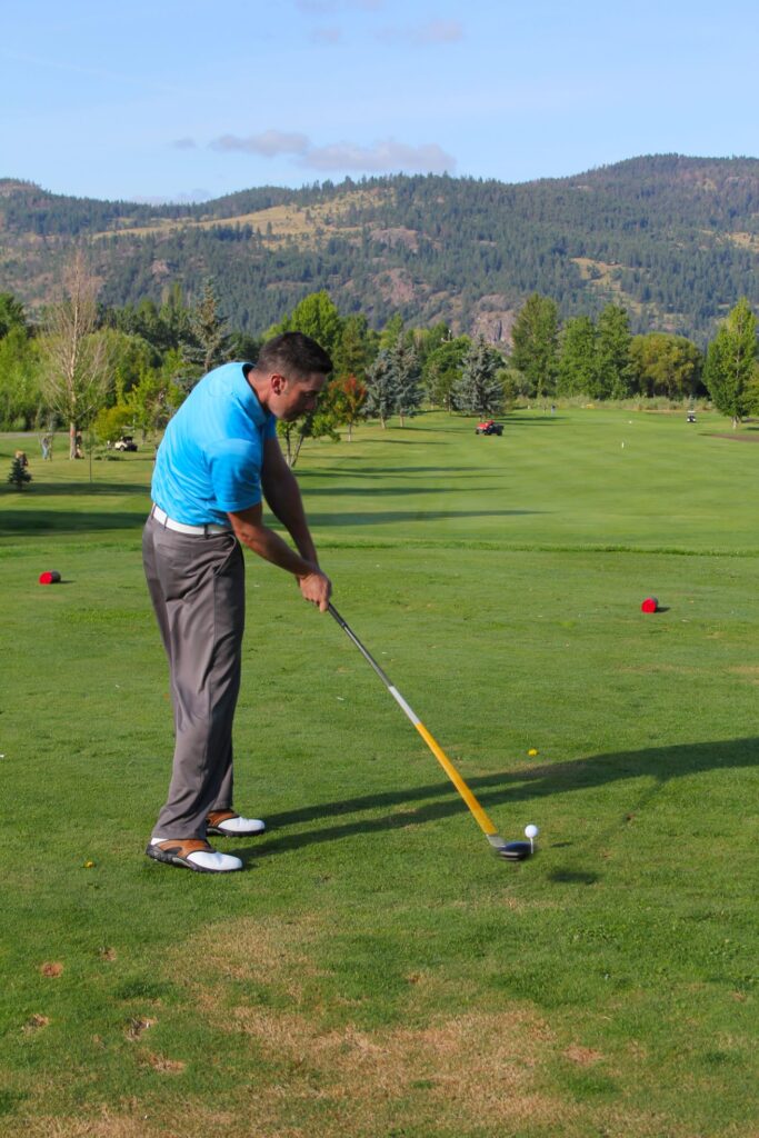 A golfer hits a drive on a golf course with mountains in the background. 